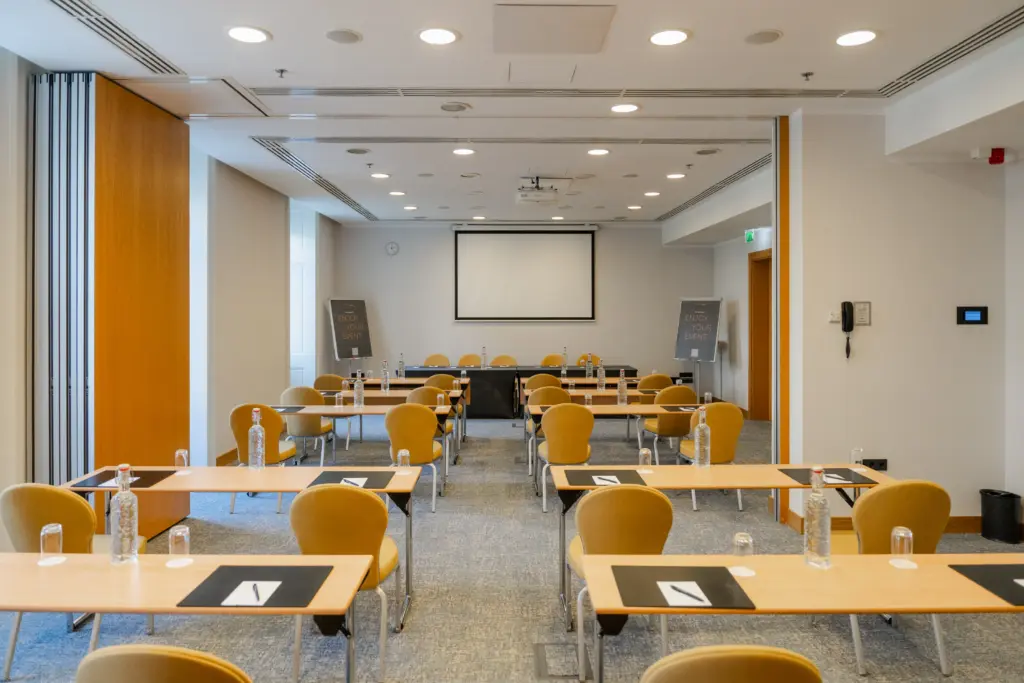 Jászai Mari konferenciaterem iskolapados elrendezésben a Courtyard by Marriott Budapest City Centerben, palackozott vízzel és jegyzetes asztalokkal.