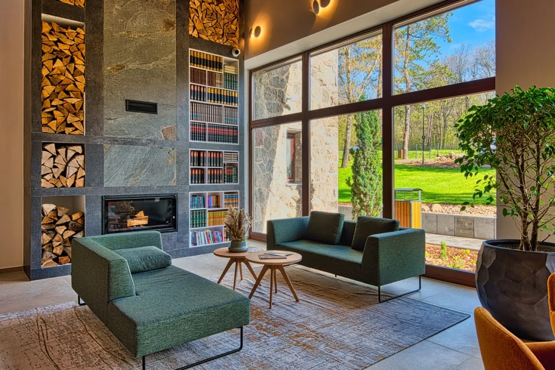 A Cardoner Hotel Dobogókő lobby pihenősarkában fatüzelésű kandalló, könyvespolc, zöld kanapék és panorámaablak látható az ősfás parkra