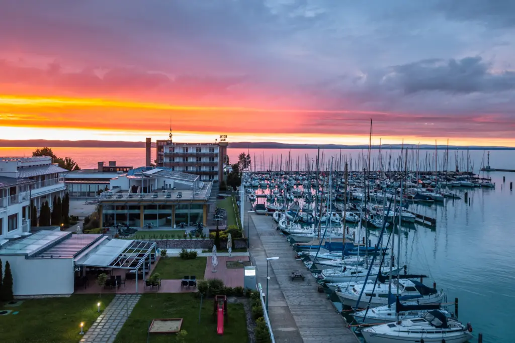 Esti légi felvétel a Hotel Yacht Siófokról rózsaszín-narancssárga égbolttal, ahol a vitorláskikötő és a Balaton látható naplementében