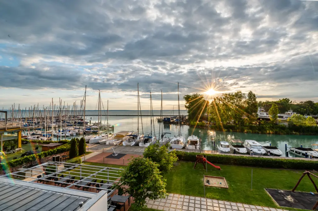 A Hotel Yacht Siófok vitorláskikötője napfelkeltében, ahol vitorlások, yachtok és a Balaton látható arany fényben