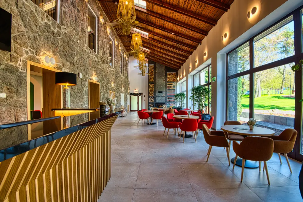 A Cardoner Hotel Dobogókő lobby-ja, ahol terméskő burkolat, fatüzelésű kandalló, fa gerendás mennyezet és designer csillár látható panorámaablakokkal az erdőre