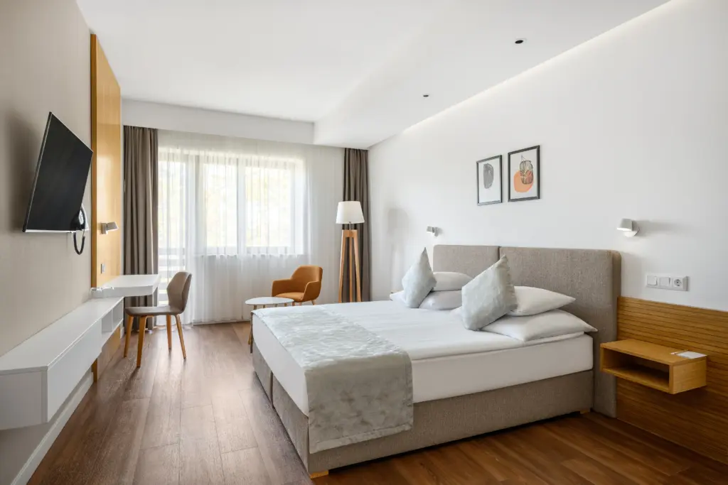 A Cardoner Hotel Dobogókő modern szobája, ahol fa padló, bézs bútorok és természetes fény látható panorámaablakon keresztül