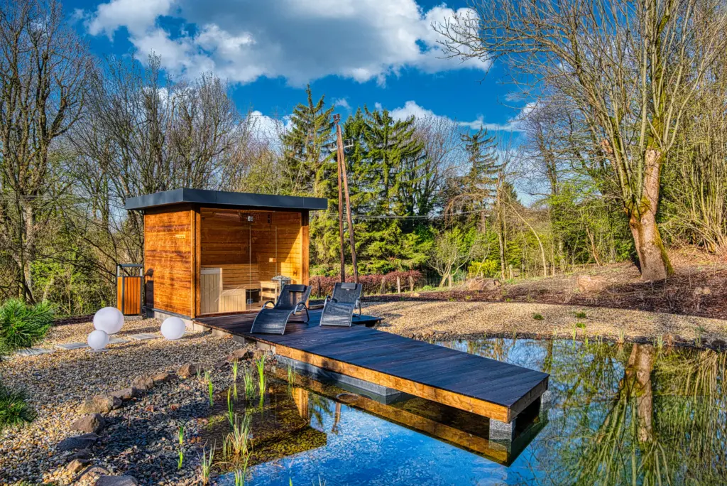 A Cardoner Hotel Dobogókő kültéri panoráma szaunája és merülődézsája, ahol fa szaunakabin és fa terasz látható kis tó mellett erdős környezetben