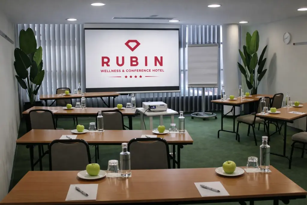 A Rubin Wellness & Conference Hotel Budapest Nefrit tárgyalóterme iskolapados elrendezéssel, ahol projektor, vetítővászon és zöld dísznövények láthatók