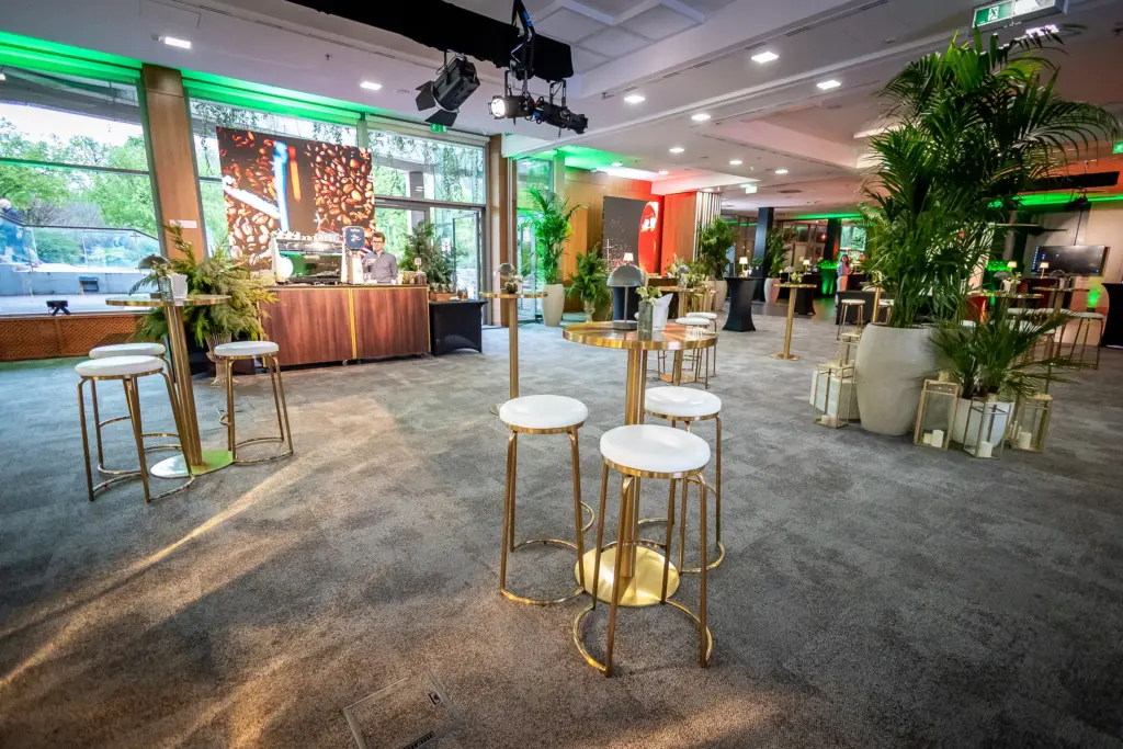A Larus Event & Catering Budapest rendezvénytere állófogadásra berendezve, ahol arany bárszékek, zöld LED fények és pálmák láthatók természetes fénnyel