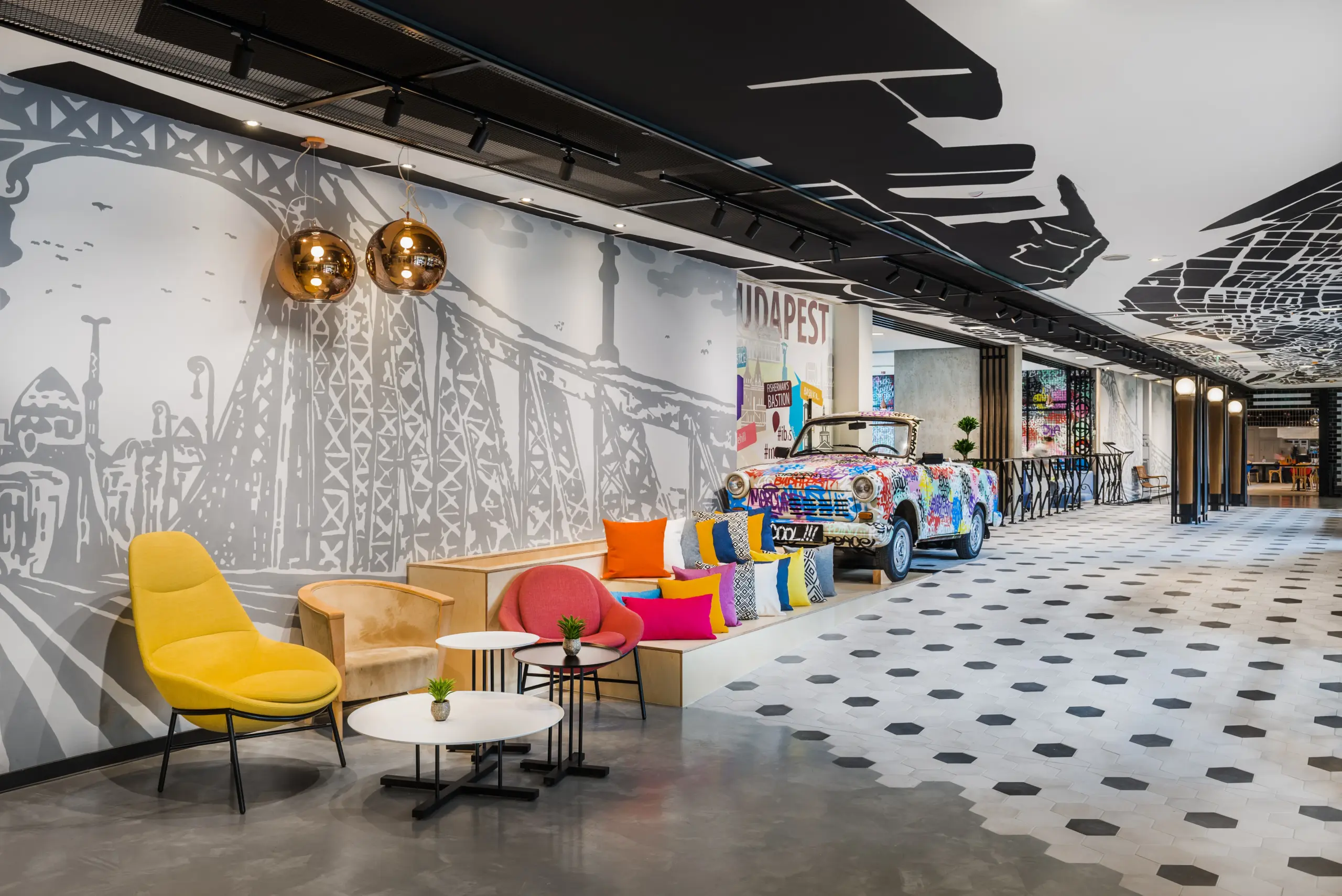 A Mercure Budapest Castle Hill lobby-ja színes Trabi autóval és Lánchíd falgrafikával, ahol sárga és rózsaszín fotelek, arany csillár és Budapest ikonjai láthatók