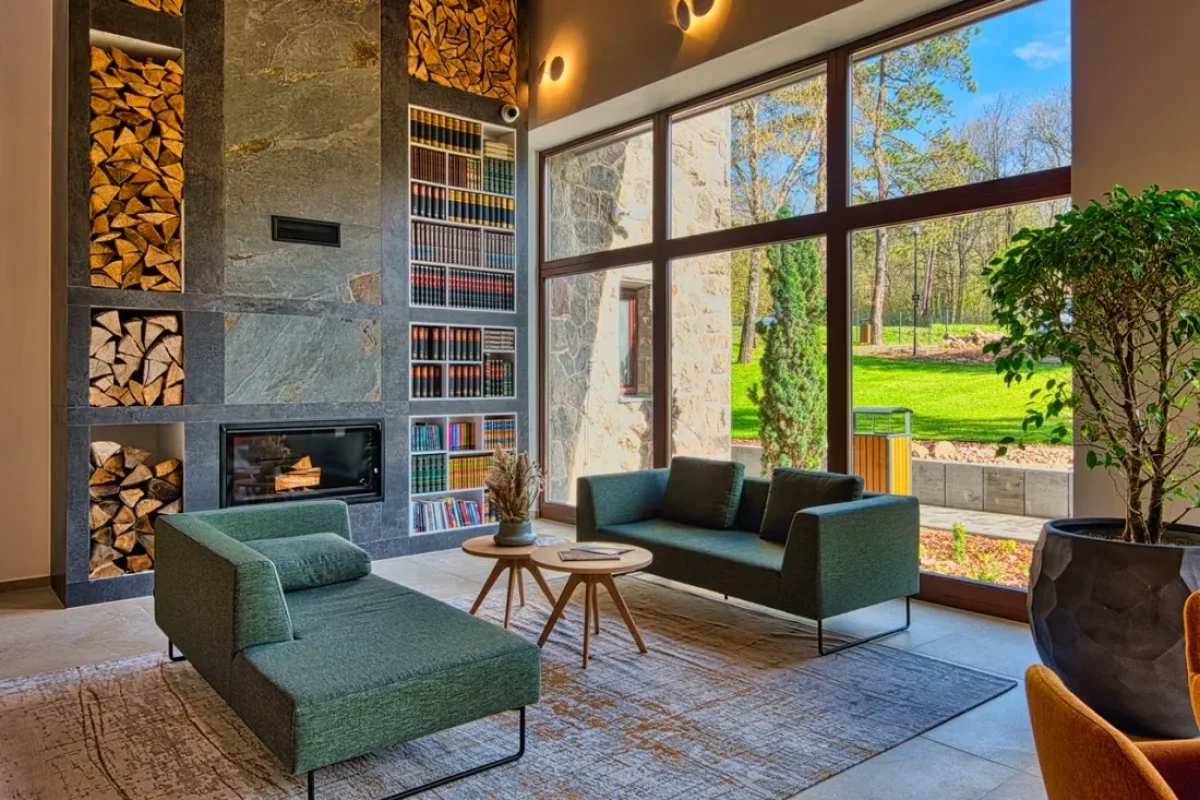 A Cardoner Hotel Dobogókő lobby pihenősarkában fatüzelésű kandalló, könyvespolc, zöld kanapék és panorámaablak látható az ősfás parkra